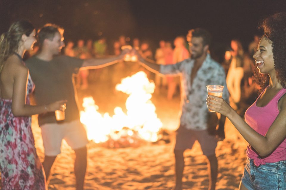 Happy friends drinking beer at beach festival with bonfire in background - Young people having fun summer vacation - Youth,holidays and party concept - Soft focus on afro girl hand glass - Warm filter